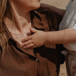 Close-up of a woman wearing a beaded necklace that spells 'MAMA' as a small child's sand-dusted hand rests on her chest/shoulder area.