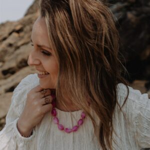 Smiling woman with long brown hair wearing a white textured blouse and pink bead necklace, standing near rocky terrain.