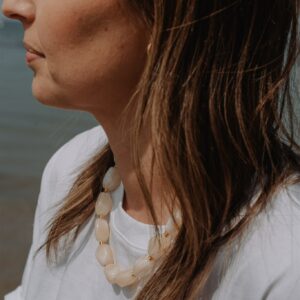 Profile of a woman facing left, wearing a cream beaded necklace and a white top by the seaside.