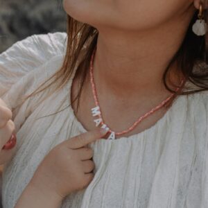 Close-up of a woman wearing a pink beaded necklace and a white textured blouse, touching the necklace at her chest with a gold earring visible.