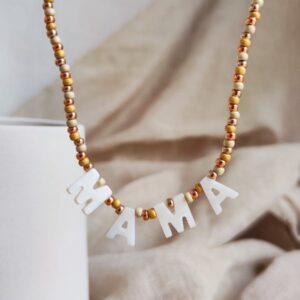 Beaded necklace with white letter charms spelling MAMA against a beige fabric backdrop in warm tones.