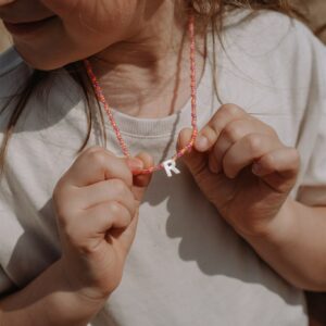 Close-up of a young girl wearing a pink beaded necklace with a white letter 'R', hair partly over her face.