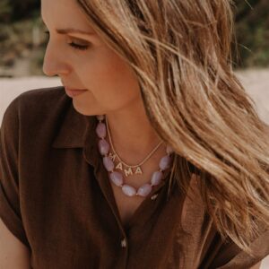 Close-up of a woman in a brown blouse wearing a pink bead necklace with a gold 'MAMA' pendant, outdoors in sunlight.