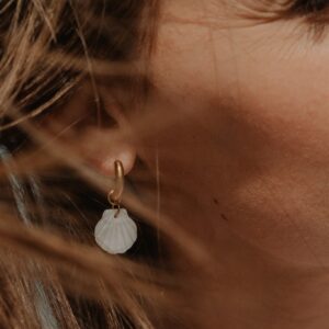 Close-up of a woman's left ear with a small gold hoop and a white shell pendant earring, hair around the ear.