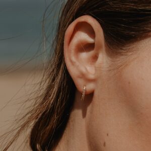 Close-up of a woman's ear wearing a small gold hoop earring with stones, hair strands nearby, skin visible on neck.