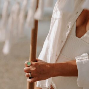 Close-up of a person in a white outfit gripping a wooden pole, wearing a green ring on their finger with a silver band nearby.