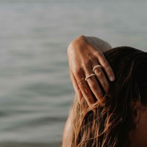 Close-up of a person with long wet hair resting a hand on their head by the sea, beads visible on rings and bracelet.
