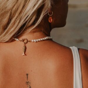 Back view of a woman with blonde hair, wearing a pearl necklace and gold hoop earring with a red pendant; a vertical tattoo runs along the spine visible on the back.