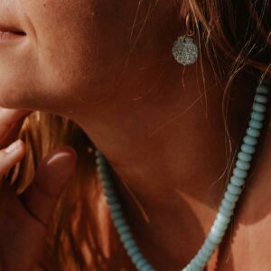 Close-up of a woman's ear with a textured silver disc earring and a turquoise bead necklace in warm sunlight.