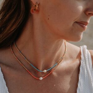 Close-up of a woman's neck and chin, showing layered coral and blue beaded necklaces with a small white shell bead; gold hoop earring with a pink flower pendant visible.