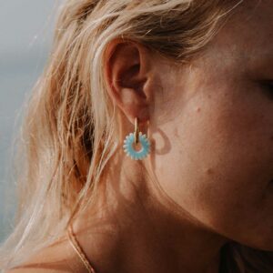 Close-up of a woman's ear with a turquoise flower-shaped earring, blonde hair framing the ear, and a thin multicolored necklace visible on the neck.