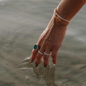 Hand with pearl bracelets and turquoise ring dipping into calm water near shore.