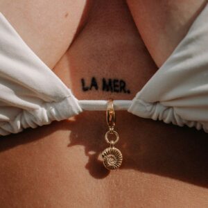 Close-up of a gold shell pendant necklace hanging between white fabric against sun-kissed skin, with the tattoo 'LA MER' visible nearby.