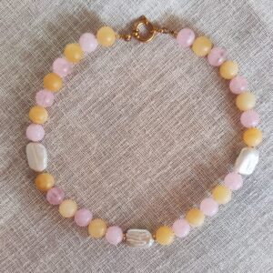 Beaded bracelet with pink and yellow round beads and three white rectangular beads, gold clasp, on beige fabric.