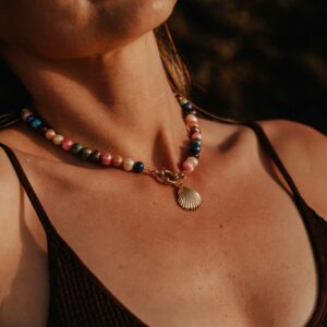 Close-up of a multicolored beaded necklace with a shell pendant resting on the chest above a brown tank top.