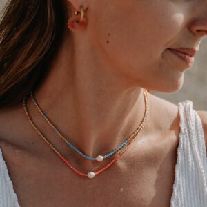 Close-up of a woman's neck wearing layered beaded necklaces in coral, turquoise, and beige tones, with an orange flower hoop earring visible on the ear.