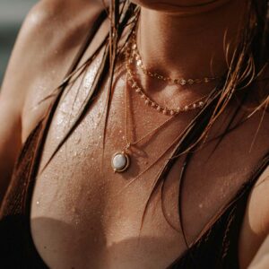 Close-up of a woman wearing layered gold necklaces on a wet brown swimsuit, with water droplets on her skin and strands of hair framing the chest area.