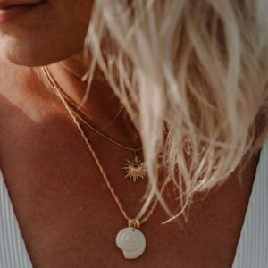Close-up of a woman’s chest with layered necklaces: a sun-shaped pendant and a white spiral shell pendant, hair partially covering the face.