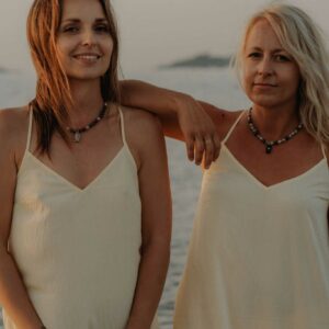 Two women in white dresses stand close together by the water, smiling at the camera during sunset.