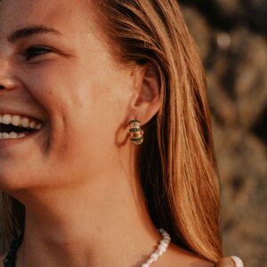 Close-up of a smiling woman's face in warm sunlight, showing green beaded earrings and a layered pearl necklace.