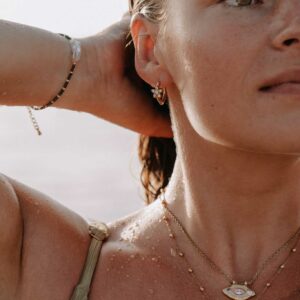 Close-up of a person’s ear and neck wearing layered gold shell jewelry on a sunlit beach, with water droplets on skin.