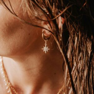 Close-up of a woman's ear wearing a gold hoop earring with a star pendant, with wet hair and layered necklaces in the background.