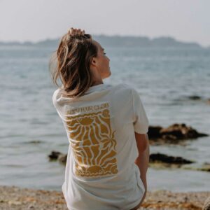 Person sitting on rocky shore by the sea, facing away, wearing a white T‑shirt with a yellow sun graphic.