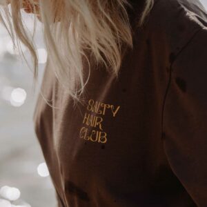 Close-up of a person with messy blond hair obscuring their face, wearing a brown shirt with gold text reading 'SALTY HAIR CLUB' against a bright, bokeh background.