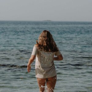 Woman with shoulder-length hair in a light patterned dress stands in shallow ocean water with waves splashing around her legs at the shore.